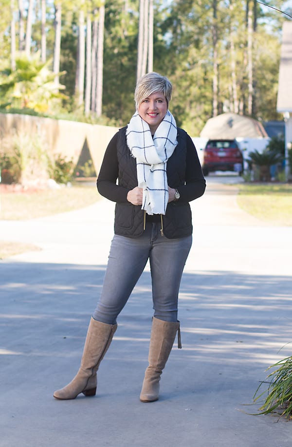Over 40 fashion blogger in winter outfit featuring grey jeans, black sweater, black vest and tan boots.